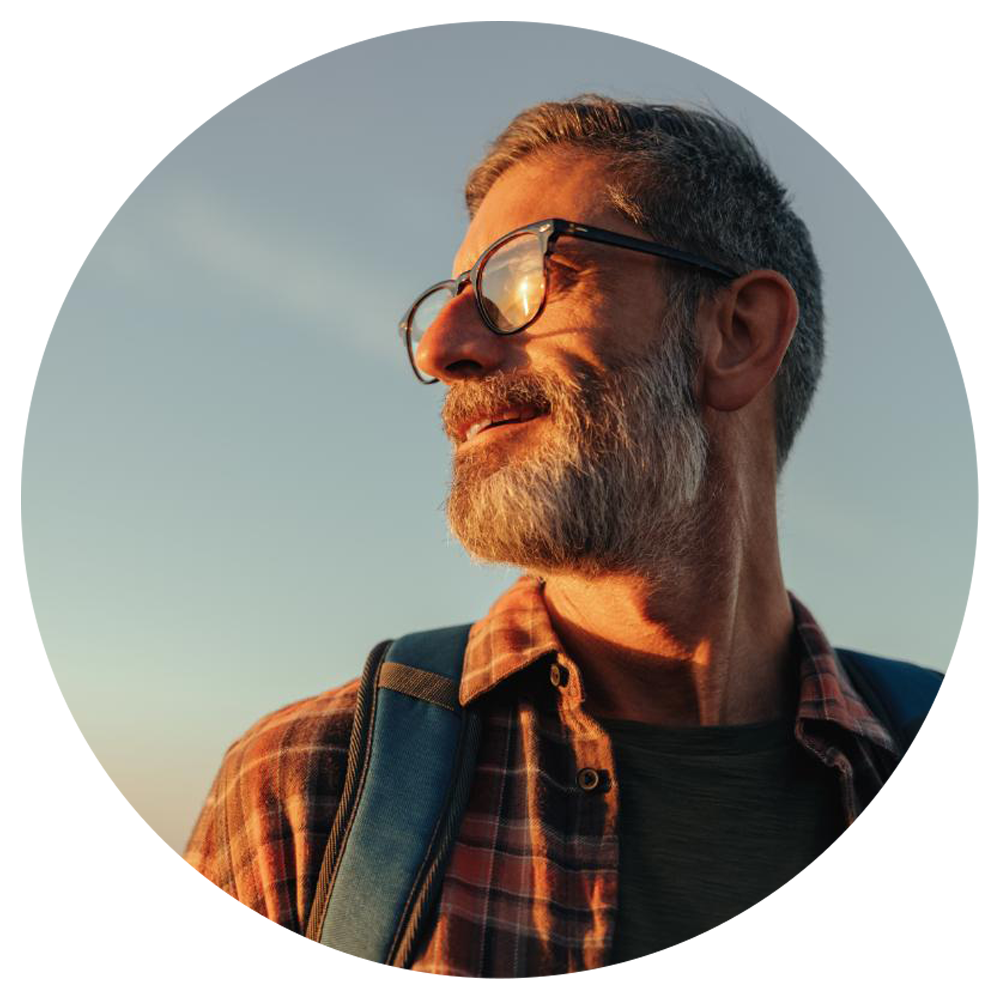 an older man with glasses and wearing a backpack looks to his side at the sunset