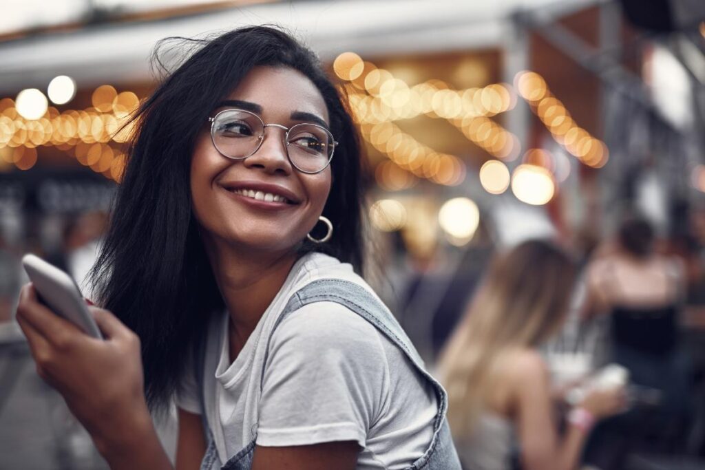 a photo of a woman with glasses with has her phone in her hand and smiling off into the distance