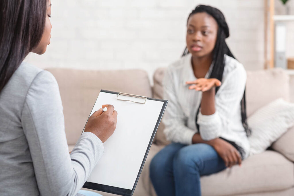 a woman sits on a couch talking to the woman across from her who is taking notes on her clipboard