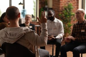 A man speaks to a small therapy group sitting in a circle, sharing his thoughts while others listen attentively in a brick-walled room.