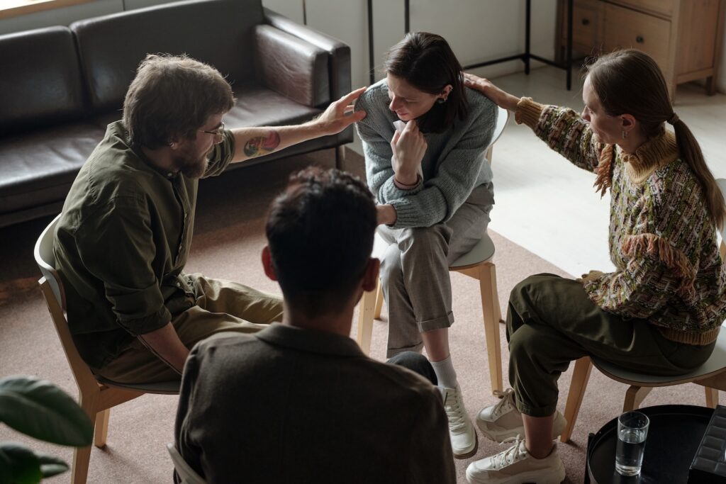 A support group comforts a woman who appears emotional, with two people placing reassuring hands on her shoulders.