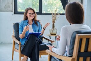 A smiling therapist with glasses holds a clipboard and talks with a client in a bright, comfortable office.
