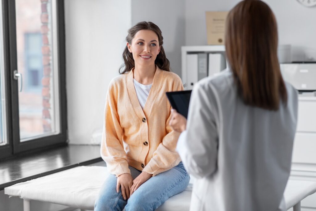 A woman sits on an exam table smiling and listening to a healthcare professional holding a tablet in a medical office.