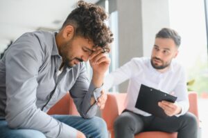 Man sitting with his head down in distress during a therapy session, while a therapist sitting nearby offers comfort and holds a clipboard.